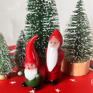 Könnte beinhalten: Zwei Weihnachtsfiguren aus Glas, die den Weihnachtsmann und einen Wichtel darstellen, mit roten Hüten und Mänteln sowie weißen Bärten. Sie stehen auf einer roten Oberfläche mit weißen Sternen, umgeben von kleinen grünen Weihnachtsbäumen. Die Santa-Figur ist etwa 5 cm groß.