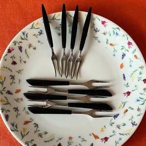 Op de afbeelding: Een set van zes cocktailvorken met zwarte handvatten op een wit bord met een bloemenpatroon. De vorken zijn in een waaier vorm gerangschikt met de handvatten die naar het midden van het bord wijzen.