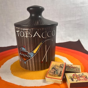 May include: A brown tobacco jar with a black lid and the word "TOBACCO" in white. The jar features a painted pipe design. Three vintage matchboxes are in front of the jar, with colourful illustrations.