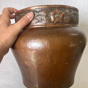 Könnte beinhalten: Eine gehämmerten Kupfervase mit breitem Korpus und dekorativem Blumen- und Blätterrand. Die Vase hat eine warme, rotbraune Farbe und eine strukturierte Oberfläche. Der obere Rand zeigt geprägte Blumen und Blätter.