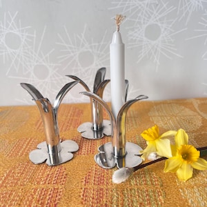 May include: A set of three silver-colored metal candle holders with a flower-like design. One holder has a white candle. Yellow daffodils and a sprig of pussy willow are on a textured orange and yellow surface.