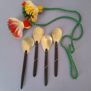 May include: A collection of five decorative spoons with silver-colored, teardrop-shaped bowls and black handles. The handles have silver accents. Two colorful tassels with green rope are at the top of the image, with red, yellow, and blue accents.