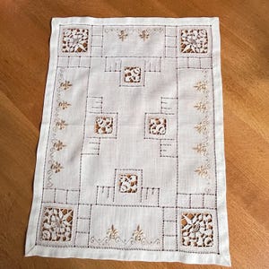 May include: Cream-colored rectangular doily with a geometric design. The doily features embroidered squares with openwork lace centers, connected by straight lines and dotted borders. Floral and leaf motifs adorn the edges and corners, creating a delicate, vintage aesthetic.