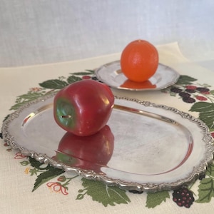 Può includere: Una mela rossa con un gambo verde poggia su un vassoio ovale d'argento con un bordo decorativo. Un'arancia è su un piatto d'argento più piccolo e abbinato sullo sfondo. Gli oggetti sono su una tovaglia a motivi floreali.