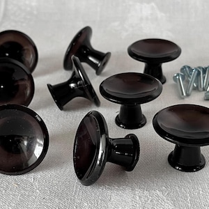 May include: A set of ten black cabinet knobs with a round, slightly concave top and a short, cylindrical base. The knobs are arranged in a semi-circle on a white surface.