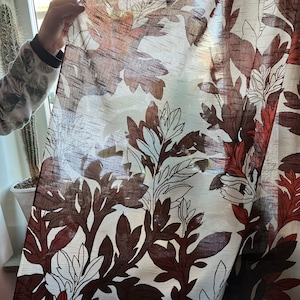Puede incluir: Una cortina blanca transparente con un patrón floral marrón y blanco. La cortina tiene un aspecto texturizado, tejido y presenta hojas y flores grandes y estilizadas.