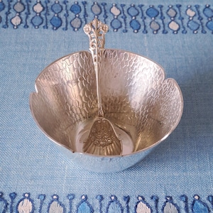 Può includere: Una ciotola da portata color argento con un cucchiaio abbinato. La ciotola ha una superficie strutturata e un bordo smerlato. Il cucchiaio è all'interno della ciotola. Il set è su una superficie blu fantasia.