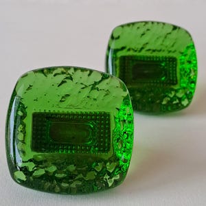 May include: Two square, green glass cabinet knobs. Each knob has a textured surface and a rectangular, recessed design in the centre. The knobs are a vibrant, transparent green, and appear to be handmade.