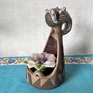 May include: A ceramic candle holder shaped like a woman with a flower design. The holder is brown with white and black accents. The flower is pink and white with green leaves.