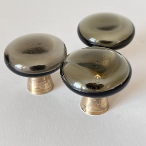 May include: Three gray glass cabinet knobs with brass bases are arranged on a white surface. The knobs have a rounded, mushroom-like shape with a dark gray, reflective finish. The brass bases add a touch of warmth.