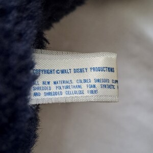 Puede incluir: Una etiqueta blanca con texto azul que dice "COPYRIGHT &copy; WALT DISNEY PRODUCTIONS ALL NEW MATERIALS, COLORED SHREDDED CLIP SHREDDED POLYURETHANE FOAM SYNTHETE AND SHREDDED CELLULOSE FIBERS".