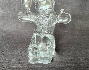 May include: A clear glass Santa Claus figurine with a detailed beard, hat, and outstretched arms. The Christmas decoration is sitting down and has a textured surface. The figurine is a festive holiday decoration.