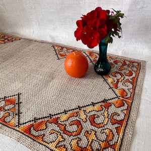 May include: A beige linen table runner with an embroidered orange and brown geometric design. A small blue vase holds red flowers. An orange sits on the runner.