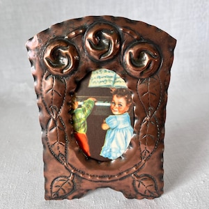 Può includere: Una cornice color rame con un design decorativo a foglie e volute. La cornice contiene un'immagine vintage di due bambini, uno con un vestito blu, l'altro con una camicia a quadri verdi, vicino a un pianoforte. La cornice è alta circa 13 cm.