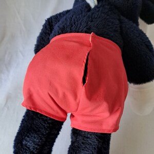 Puede incluir: Un juguete de peluche negro con pantalones cortos rojos. Los pantalones cortos tienen una costura en la parte posterior.
