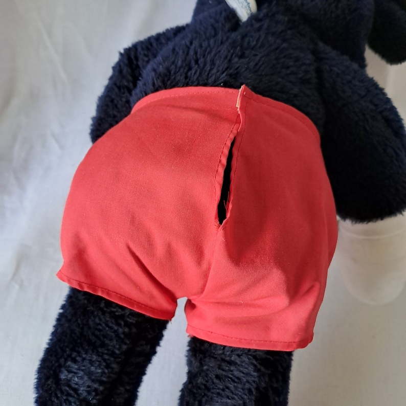 Puede incluir: Un juguete de peluche negro con pantalones cortos rojos. Los pantalones cortos tienen una costura en la parte posterior.