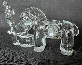 May include: A clear glass candle holder in the shape of a horse with a rider. The horse has a textured mane and a saddle-like area to hold a candle. The rider is seated and appears to be playing a drum. The candle holder is on a black surface.