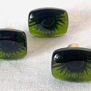 May include: Three decorative glass bottle stoppers. Each stopper is square with a dark green top and a light green, eye-like design. The stoppers have a light wood base. The stoppers are on a white surface.