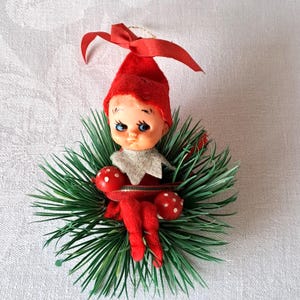 Op de afbeelding: Een vintage kerstornament met een popachtige elf met een rode hoed en outfit, genesteld in groene dennennaalden. De elf heeft blauwe ogen en houdt twee rode paddenstoelen met witte stippen vast. Bovenop zit een rode strik.