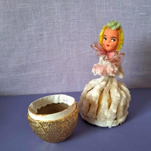 Puede incluir: Adorno vintage con forma de muñeca con cabeza amarilla y verde, lazo rosa y vestido de piel blanca. Junto a él, un recipiente dorado con textura de huevo y un interior blanco. Los artículos están sobre una superficie púrpura.