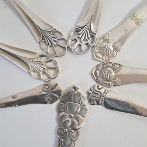Puede incluir: Vista aérea de cubiertos antiguos dispuestos en un patrón circular. Las cucharas y tenedores de plata tienen diseños florales y de volutas ornamentados en los mangos. La cubertería está sobre un fondo blanco.
