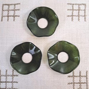 May include: Three dark green, irregularly shaped glass coasters with a central hole are arranged on a white fabric surface. The coasters have a glossy finish and a textured appearance. The background features embroidered cross-stitch patterns.