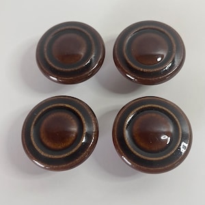 Op de afbeelding: Vier bruine keramische kastknoppen met een glanzende afwerking. Elke knop heeft een cirkelvormig ontwerp met concentrische ringen en een centrale, iets verhoogde cirkel. De knoppen zijn gelijkmatig verdeeld tegen een witte achtergrond.