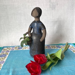 May include: A ceramic figure of a woman in a blue dress, holding a basket with a plant. The figure is dark blue and brown. Two red tulips with green stems and leaves are in the foreground. The figure is on a blue and white patterned surface.