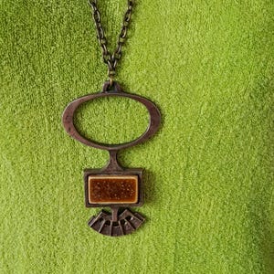 Peut inclure: Un collier pendentif vintage avec une chaîne et un pendentif de couleur bronze. Le pendentif a un dessus ovale, un centre rectangulaire avec un motif tacheté marron et un bas en forme d'éventail. Le collier est présenté sur une surface verte texturée.