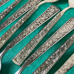 Puede incluir: Primer plano de varias cucharas plateadas con intrincados diseños florales en los mangos. Las cucharas están dispuestas en diagonal, mostrando el diseño detallado. Las cucharas están sobre una superficie verde.