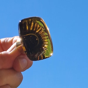 Op de afbeelding: Een rechthoekige, amberkleurige kastknop met een goudkleurige basis. De knop heeft een ingewikkeld, op een zonnestraal lijkend ontwerp op het oppervlak, dat licht reflecteert. De achtergrond is een heldere, helderblauwe lucht.