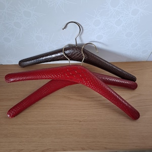 May include: Three padded clothing hangers. One is red, and two are brown. The hangers have a textured, snakeskin-like pattern. The hooks are silver and gold-toned. The hangers are on a wooden surface.