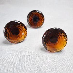 May include: Three decorative glass knobs with a swirling, textured design. The knobs are a mix of amber and dark brown, with a glossy finish. They are round and appear to be handmade, suitable for furniture or cabinet hardware.