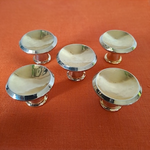 May include: Five gold-toned cabinet knobs with a polished finish. Each knob features a rounded, flat top with a silver-colored rim and a short, cylindrical base. The knobs are arranged on an orange surface.