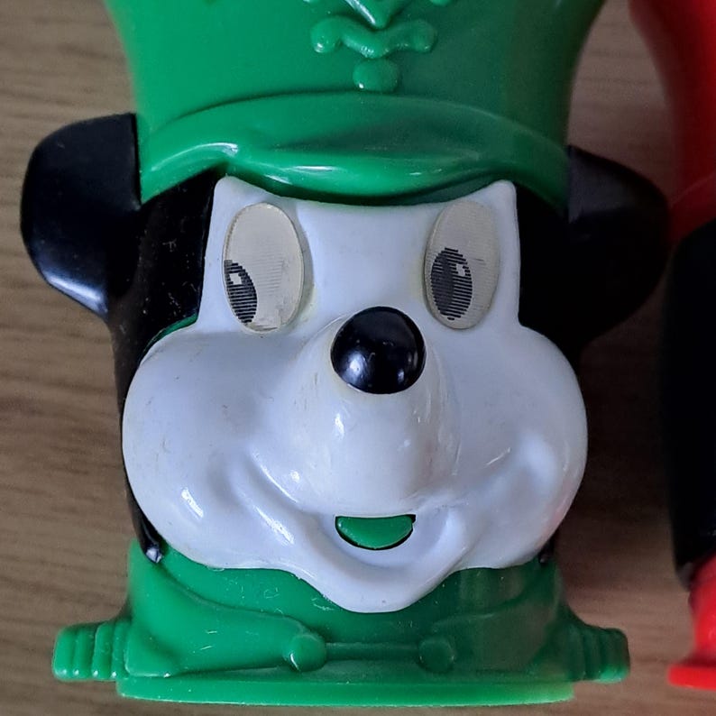 May include: Close-up of a vintage Mickey Mouse toy figure. The figure has a white face, black ears, and a green hat and collar. The figure's eyes are oval-shaped with a black nose and a smiling mouth. The figure is made of plastic.