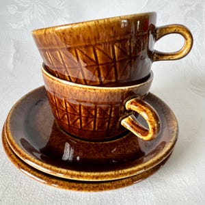 May include: Two brown ceramic teacups with a geometric pattern stacked on top of each other on a brown saucer.