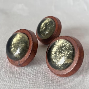 Op de afbeelding: Drie decoratieve houten knoppen met een helder, gewelfd midden gevuld met een glinsterend, goudgroen materiaal. De knoppen hebben een rijke, roodbruine houten afwerking, wat een contrast creëert met de lichtgevende centra. Ideaal voor meubels of knutselprojecten.