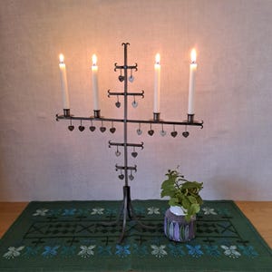 Op de afbeelding: Een smeedijzeren kandelaar met vier brandende kaarsen. De kandelaar heeft een centrale verticale steun met horizontale armen die de kaarsen vasthouden. Kleine hartvormige accenten hangen aan de armen. Een groen geborduurde tafelloper en een kleine vaas met groen staan op de voorgrond.