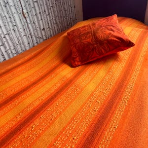 May include: An orange and yellow striped bedspread with a matching orange pillow. The pillow has a floral design in a darker shade of orange. The bedspread has a textured, woven appearance.