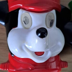 May include: Close-up of a vintage Mickey Mouse figurine. The figurine features a white face, black ears, and a red hat and scarf. The eyes are oval-shaped with a detailed iris. The nose is a glossy black sphere, and the mouth is a small red slit.