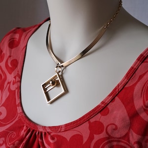 May include: Gold-coloured necklace with a geometric pendant. The pendant has a square frame and a sphere accent. The necklace has a flat, choker-style band and a chain. The pendant is approx. 3.8 cm wide.