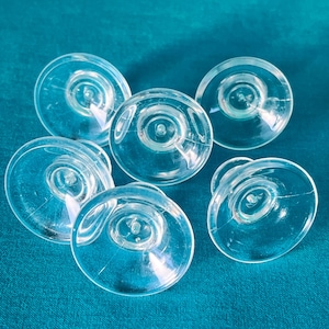 Op de afbeelding: Zes transparante zuignapjes van plastic met een ronde basis en een kleine centrale steel. De zuignapjes zijn in een halve cirkel op een blauw oppervlak gerangschikt.
