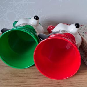 May include: Two ceramic bell-shaped figurines with white faces, black eyes, and black hats. One bell is green, the other is red. The figurines are lying on a wooden surface.
