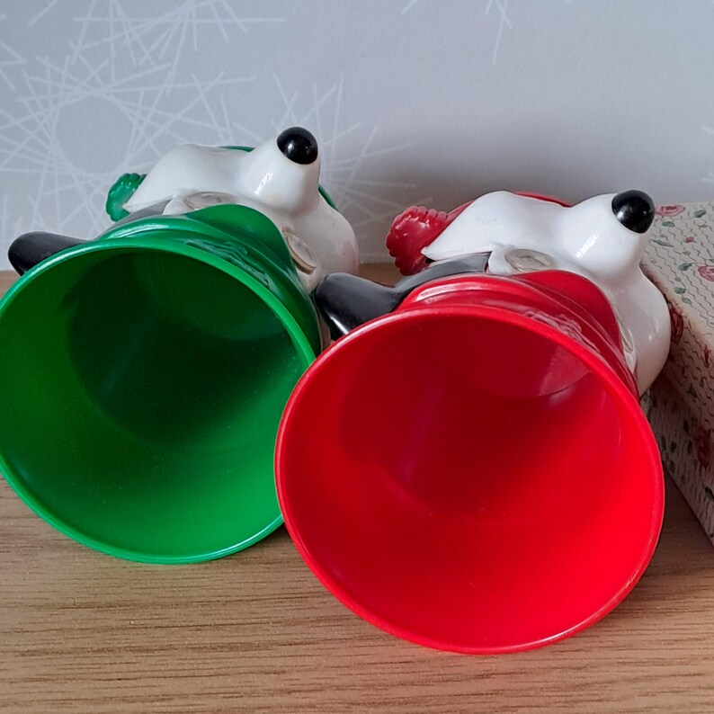 May include: Two ceramic bell-shaped figurines with white faces, black eyes, and black hats. One bell is green, the other is red. The figurines are lying on a wooden surface.