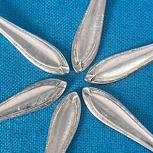 May include: Six silver spoons arranged in a circular pattern on a blue textured surface. The spoons have a decorative edge design and a polished finish, reflecting the light. The spoons are a part of a silverware set.