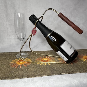 Puede incluir: Un soporte de metal dorado para botellas de vino con un asa de madera, que sostiene una botella de vino oscura. Cerca hay una flauta de champán transparente. El soporte tiene un encanto rojo decorativo. La botella está en ángulo.