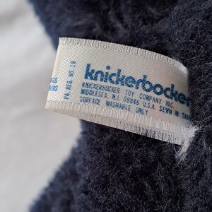 Puede incluir: Una etiqueta blanca con texto azul que dice "Knickerbocker Toy Company Inc. Middlesex NJ 08846 USA Sewn in Taiwan. PA. Reg. No. 18. Surface Washable Only."