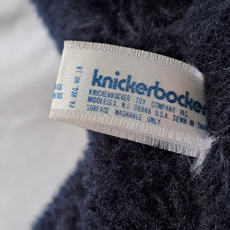 Puede incluir: Una etiqueta blanca con texto azul que dice "Knickerbocker Toy Company Inc. Middlesex NJ 08846 USA Sewn in Taiwan. PA. Reg. No. 18. Surface Washable Only."