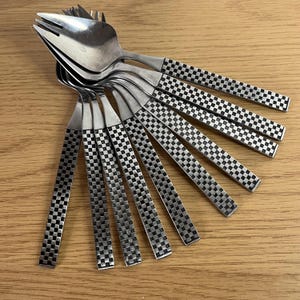May include: A set of stainless steel forks with checkered pattern handles. The forks are arranged in a fan shape, with the tines of the forks pointing upwards. The handles have a black and silver checkered design. The forks are on a light brown wooden surface.