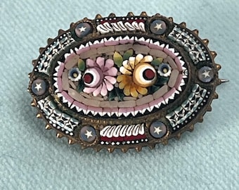 Puede incluir: Broche ovalado vintage con un diseño floral de mosaico. El broche presenta un mosaico central de flores rosas y amarillas con centros rojos, rodeado por un borde de pequeñas baldosas coloridas y detalles de estrellas. El marco de metal tiene una apariencia oscura y envejecida.